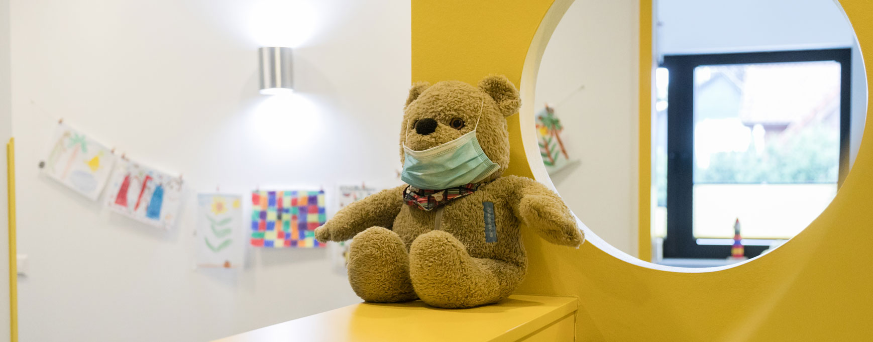 Ein großer Teddybär mit Mundschutz sint am Empfang und begrüßt die kleinen Patient_innen
