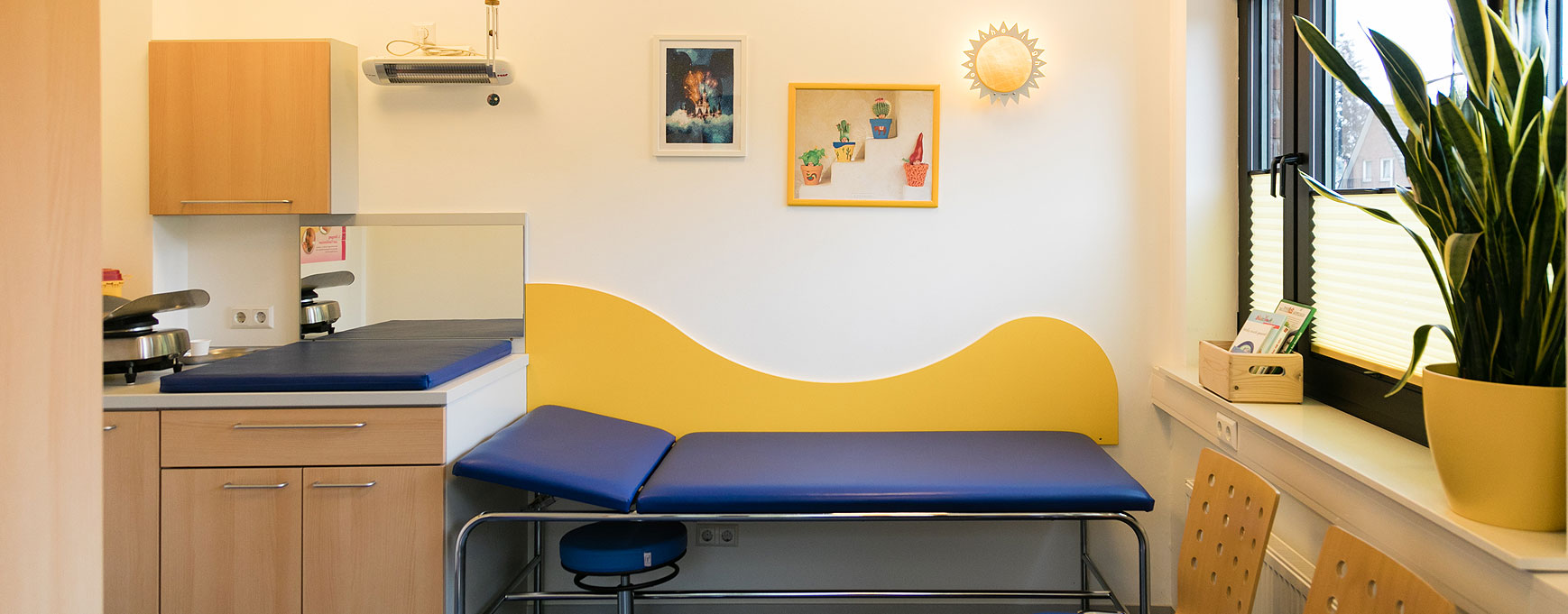 Hier siehr man ein Behandlungszimmer mit einer dunkelblauen Liege und einer gelben Holzwelle im Hintergund an der Wand.